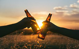 Photographie de deux personnes trinquant avec des bouteilles de bière durant un coucher de soleil.