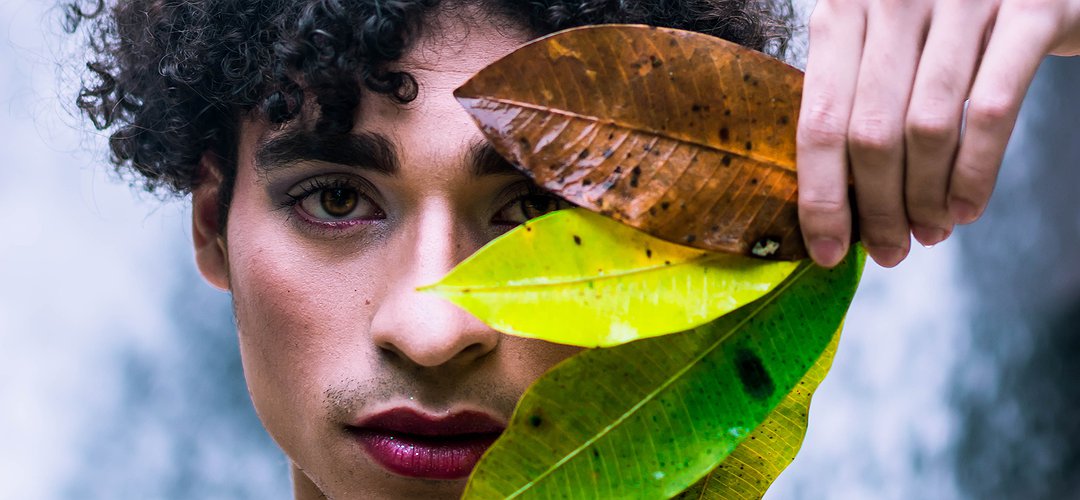 Une personne aux cheveux et sourcils noirs, rouge à lèvre et à peine de barbe tient quatre feuilles d'arbre devant son oeil gauc